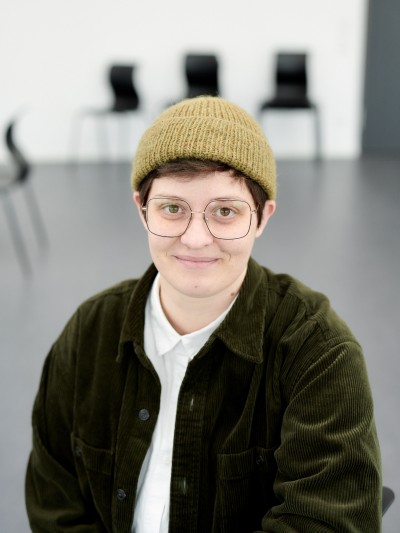 Es ist ein Potrait einer weißen Person. Der Oberkörper und der Kopf sind zu sehen sowie ein heller fast leerer Raum im Hintergrund mit grauem Boden und ein paar schwarzen Stühlen. Die Person trägt eine große Brille und eine hell-oliv-farbende Strickmütze. Unter der kurzen Mütze sind kurze dunkle Haare und beide Ohren zu sehen. Die person lächelt ganz leicht mir geschlossenem Mund. Die braunen Augen schauen direkt in die Kamera. Die person trägt ein weißes zugeknöptes Hemd und darüber eine offene dukelgrüne Cordjacke.